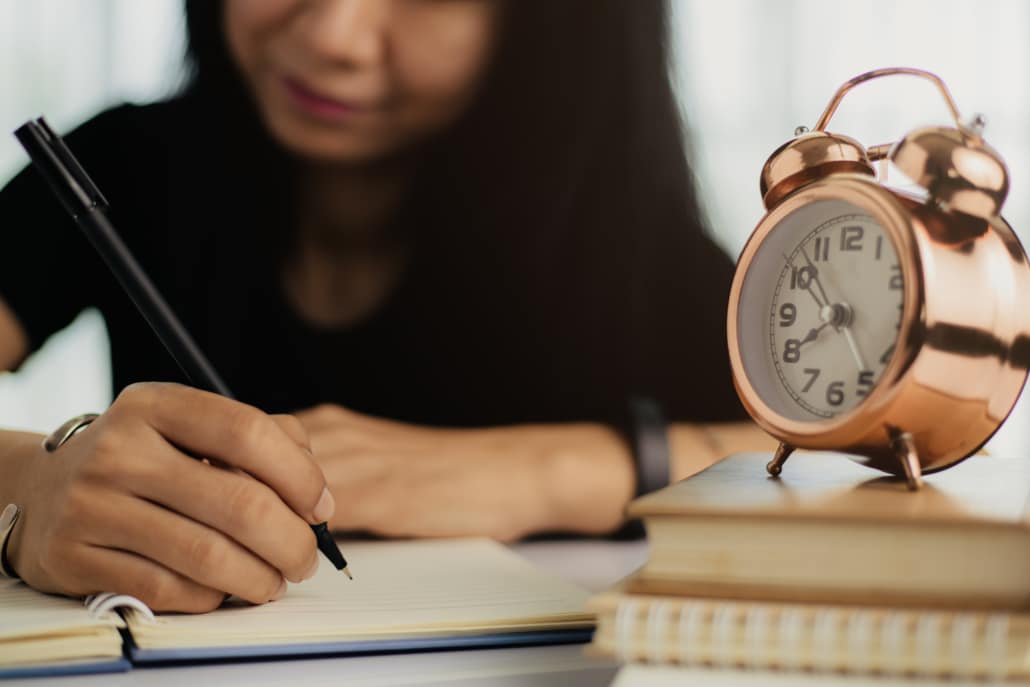 Asian woman writing on a notebook with bell alarm clock on books for study, work and time management concept